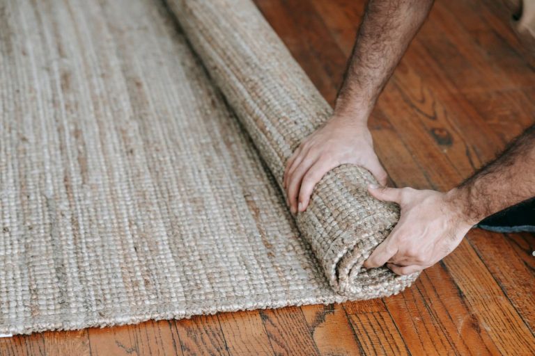 A man rolls up a rug on a wooden floor, preparing for carpet cleaning.